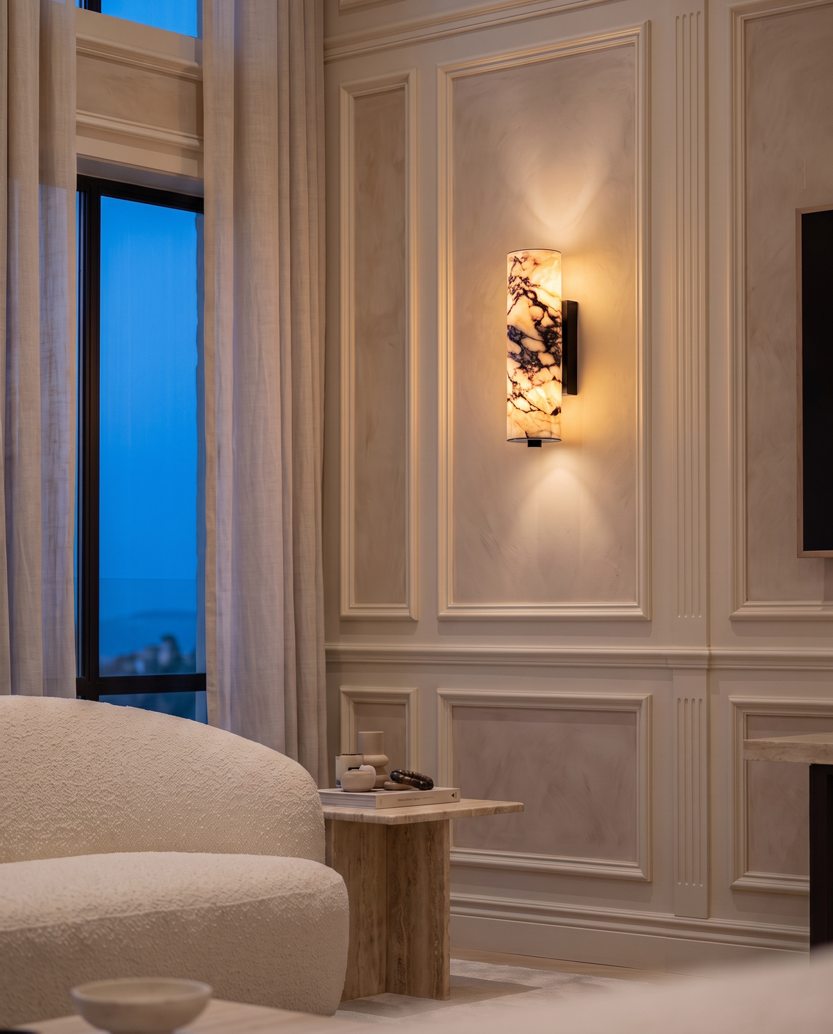 Siena marble column wall light illuminated on a panelled living room wall beside a cream sofa and tall window